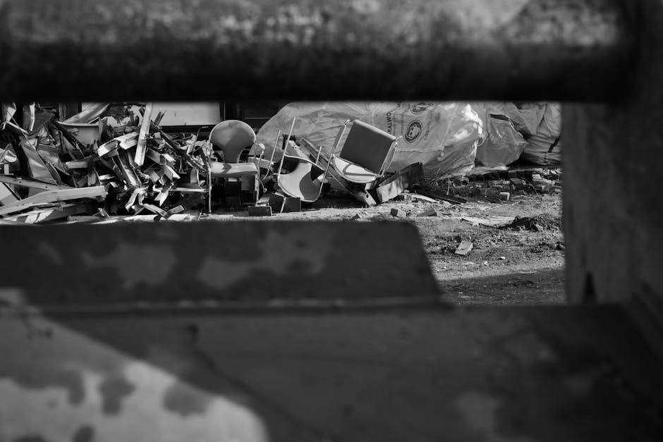 A black and white photograph displaying a pile of assorted discarded household and office waste situated outdoors on uneven ground. In the foreground, blurred metal bars partially obscure the view, su