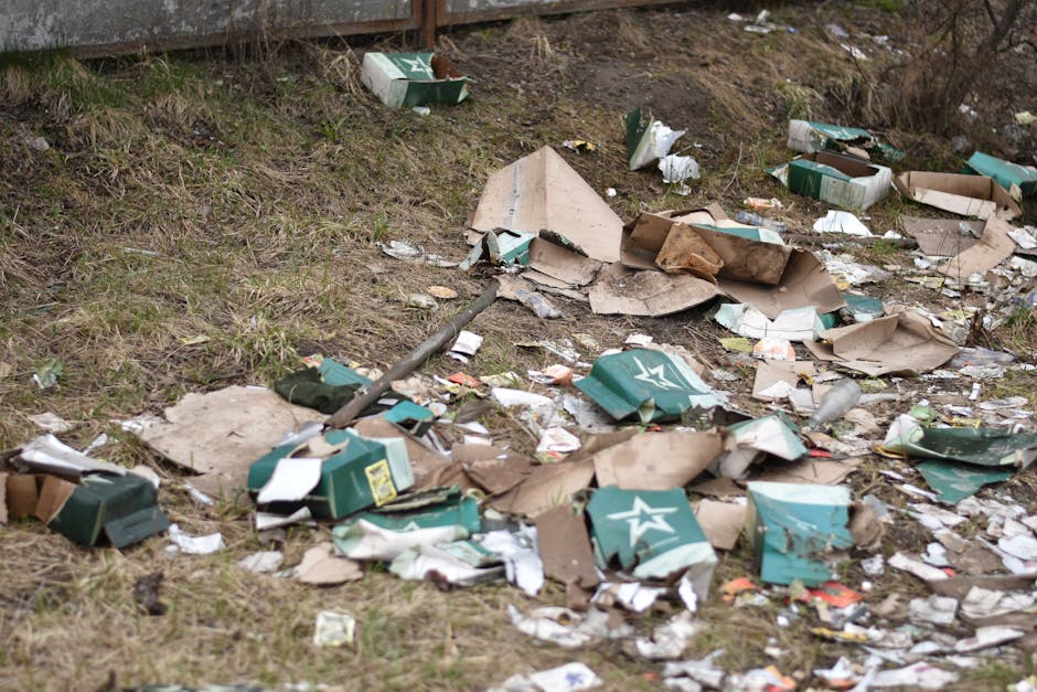 An outdoor area shows a patch of uneven ground with scattered litter, primarily consisting of flattened cardboard boxes with green and white star branding, some torn and crumpled, mixed with other pap